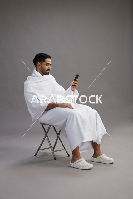 بورتريه لحاج سعودي بملابس الإحرام يستخدم الهاتف المحمول ويجلس على كرسي, الاستعداد لاداء مناسك الحج والعمره, خلفيه رماديه
