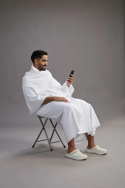 A portrait of a Saudi pilgrim in Ihram clothing using a mobile phone and sitting on a chair, preparing to perform the rituals of Hajj and Umrah, with a gray background.