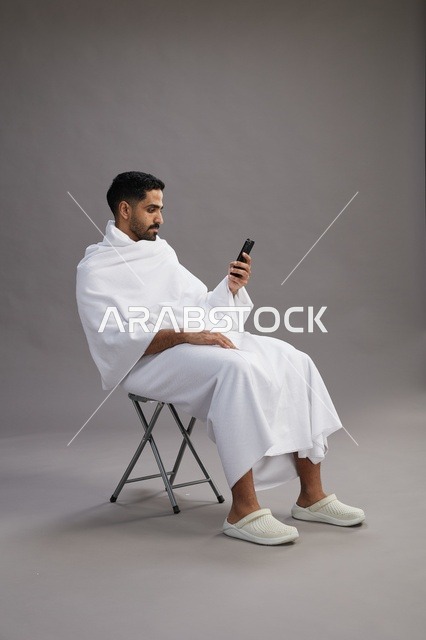 A portrait of a Saudi pilgrim in Ihram clothing using a mobile phone and sitting on a chair, preparing to perform the rituals of Hajj and Umrah, with a gray background.