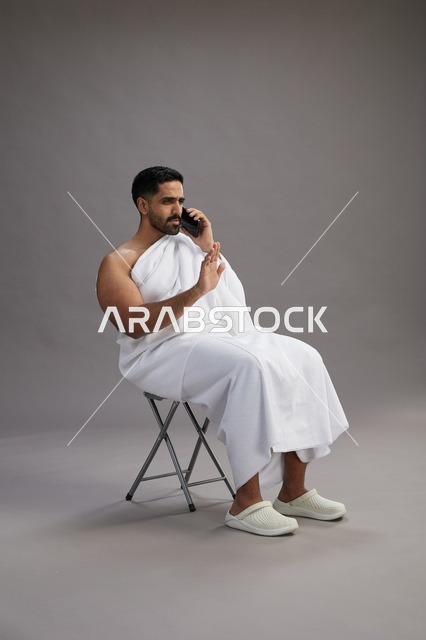 بورتريه لحاج سعودي بملابس الإحرام يستخدم الهاتف المحمول ويجلس على كرسي, الاستعداد لاداء مناسك الحج والعمره, خلفيه رماديه