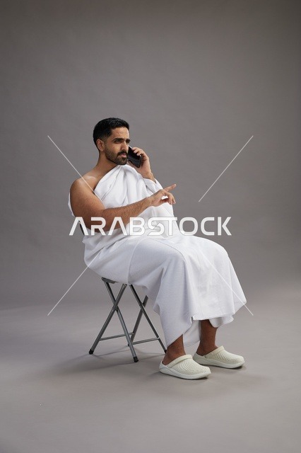 بورتريه لحاج سعودي بملابس الإحرام يستخدم الهاتف المحمول ويجلس على كرسي, الاستعداد لاداء مناسك الحج والعمره, خلفيه رماديه