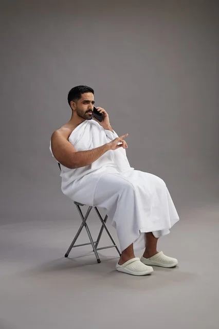 بورتريه لحاج سعودي بملابس الإحرام يستخدم الهاتف المحمول ويجلس على كرسي, الاستعداد لاداء مناسك الحج والعمره, خلفيه رماديه