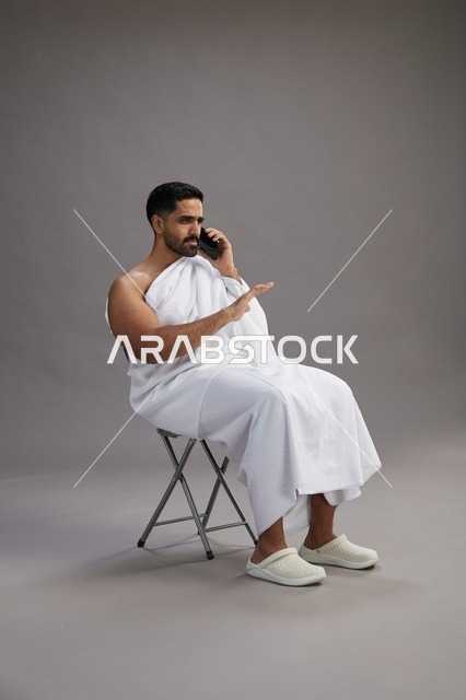 A portrait of a Saudi pilgrim in Ihram clothing using a mobile phone and sitting on a chair, preparing to perform the rituals of Hajj and Umrah, with a gray background.