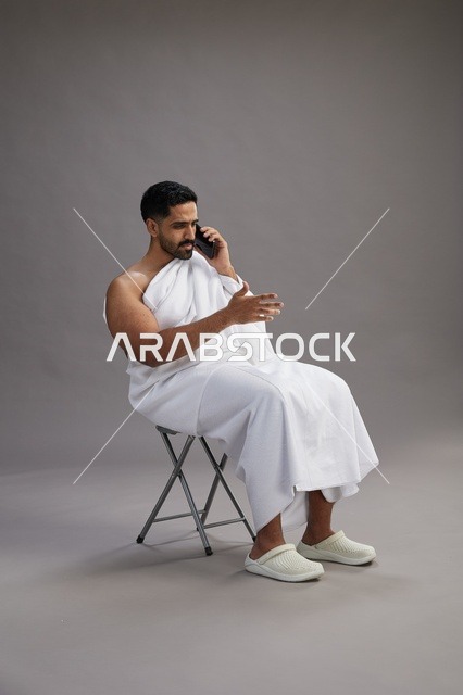 A portrait of a Saudi pilgrim in Ihram clothing using a mobile phone and sitting on a chair, preparing to perform the rituals of Hajj and Umrah, with a gray background.
