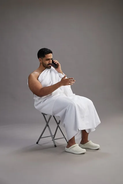 بورتريه لحاج سعودي بملابس الإحرام يستخدم الهاتف المحمول ويجلس على كرسي, الاستعداد لاداء مناسك الحج والعمره, خلفيه رماديه