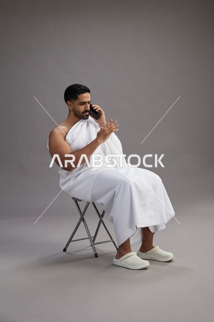 A portrait of a Saudi pilgrim in Ihram clothing using a mobile phone and sitting on a chair, preparing to perform the rituals of Hajj and Umrah, with a gray background.