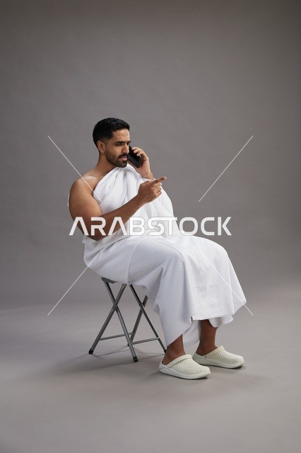 A portrait of a Saudi pilgrim in Ihram clothing using a mobile phone and sitting on a chair, preparing to perform the rituals of Hajj and Umrah, with a gray background.