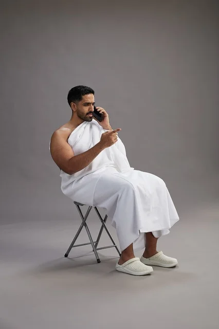 بورتريه لحاج سعودي بملابس الإحرام يستخدم الهاتف المحمول ويجلس على كرسي, الاستعداد لاداء مناسك الحج والعمره, خلفيه رماديه