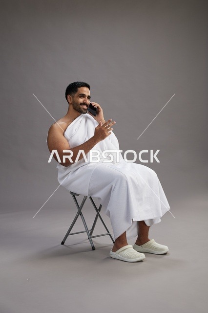 A portrait of a Saudi pilgrim in Ihram clothing using a mobile phone and sitting on a chair, preparing to perform the rituals of Hajj and Umrah, with a gray background.