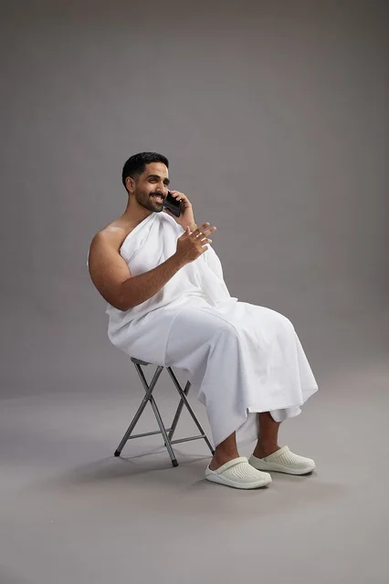 بورتريه لحاج سعودي بملابس الإحرام يستخدم الهاتف المحمول ويجلس على كرسي, الاستعداد لاداء مناسك الحج والعمره, خلفيه رماديه