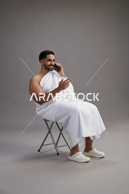 بورتريه لحاج سعودي بملابس الإحرام يستخدم الهاتف المحمول ويجلس على كرسي, الاستعداد لاداء مناسك الحج والعمره, خلفيه رماديه