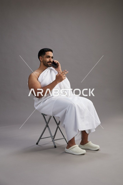 A portrait of a Saudi pilgrim in Ihram clothing using a mobile phone and sitting on a chair, preparing to perform the rituals of Hajj and Umrah, with a gray background.