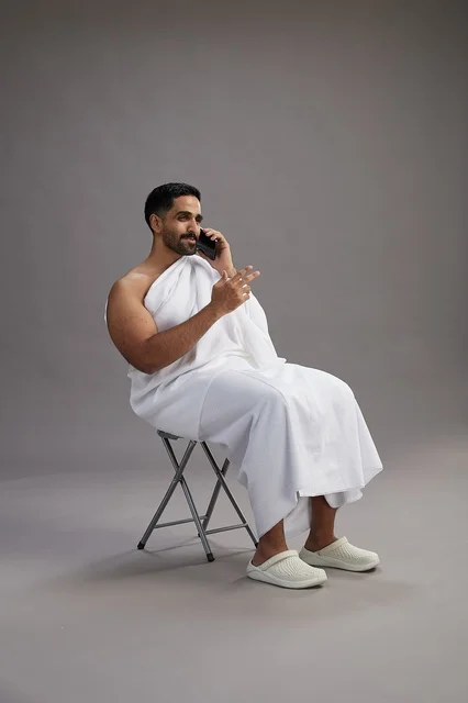 بورتريه لحاج سعودي بملابس الإحرام يستخدم الهاتف المحمول ويجلس على كرسي, الاستعداد لاداء مناسك الحج والعمره, خلفيه رماديه