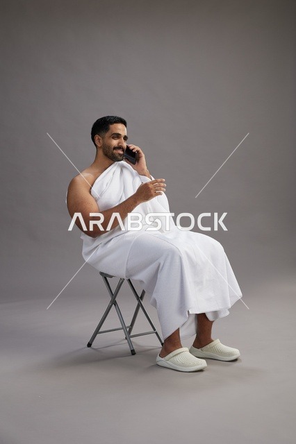 بورتريه لحاج سعودي بملابس الإحرام يستخدم الهاتف المحمول ويجلس على كرسي, الاستعداد لاداء مناسك الحج والعمره, خلفيه رماديه