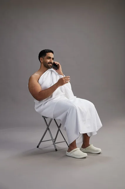 بورتريه لحاج سعودي بملابس الإحرام يستخدم الهاتف المحمول ويجلس على كرسي, الاستعداد لاداء مناسك الحج والعمره, خلفيه رماديه
