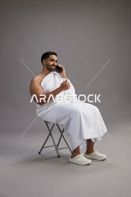 بورتريه لحاج سعودي بملابس الإحرام يستخدم الهاتف المحمول ويجلس على كرسي, الاستعداد لاداء مناسك الحج والعمره, خلفيه رماديه