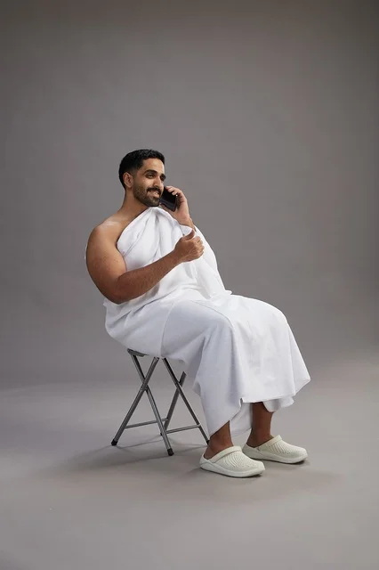 بورتريه لحاج سعودي بملابس الإحرام يستخدم الهاتف المحمول ويجلس على كرسي, الاستعداد لاداء مناسك الحج والعمره, خلفيه رماديه