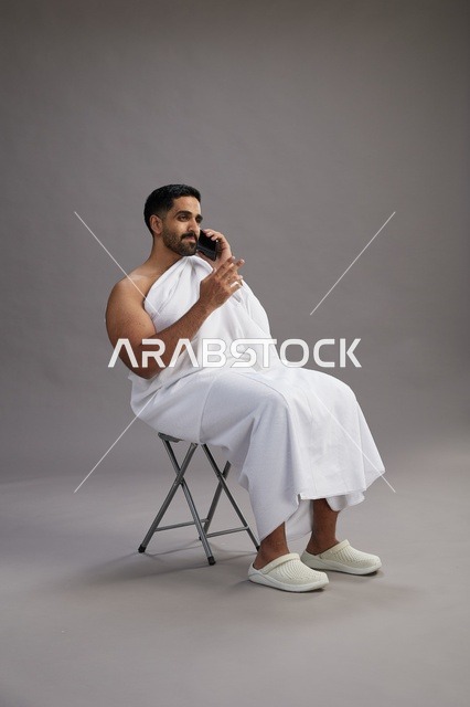 بورتريه لحاج سعودي بملابس الإحرام يستخدم الهاتف المحمول ويجلس على كرسي, الاستعداد لاداء مناسك الحج والعمره, خلفيه رماديه