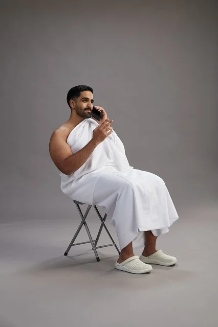 بورتريه لحاج سعودي بملابس الإحرام يستخدم الهاتف المحمول ويجلس على كرسي, الاستعداد لاداء مناسك الحج والعمره, خلفيه رماديه