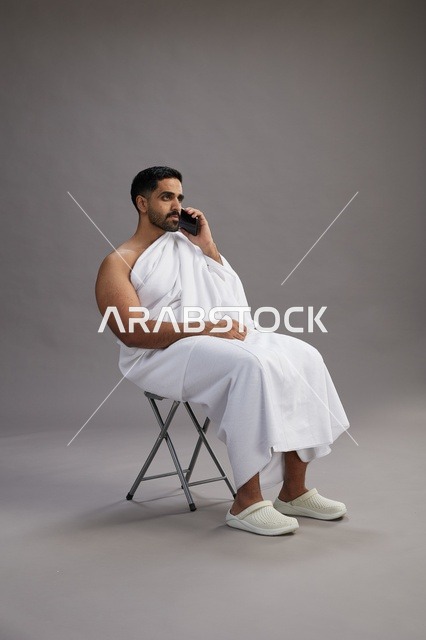 بورتريه لحاج سعودي بملابس الإحرام يستخدم الهاتف المحمول ويجلس على كرسي, الاستعداد لاداء مناسك الحج والعمره, خلفيه رماديه