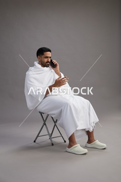 بورتريه لحاج سعودي بملابس الإحرام يستخدم الهاتف المحمول ويجلس على كرسي, الاستعداد لاداء مناسك الحج والعمره, خلفيه رماديه