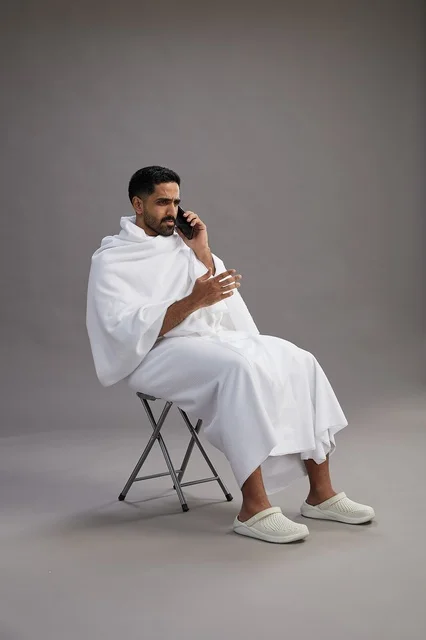 بورتريه لحاج سعودي بملابس الإحرام يستخدم الهاتف المحمول ويجلس على كرسي, الاستعداد لاداء مناسك الحج والعمره, خلفيه رماديه