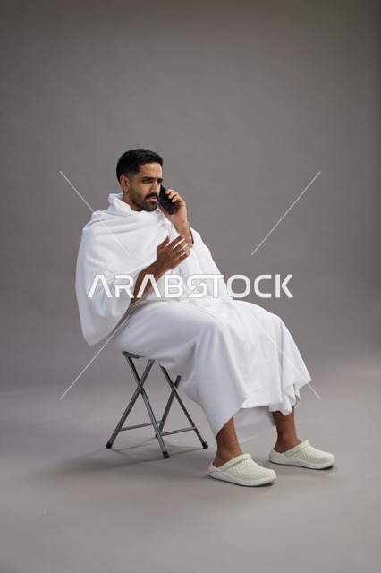 بورتريه لحاج سعودي بملابس الإحرام يستخدم الهاتف المحمول ويجلس على كرسي, الاستعداد لاداء مناسك الحج والعمره, خلفيه رماديه