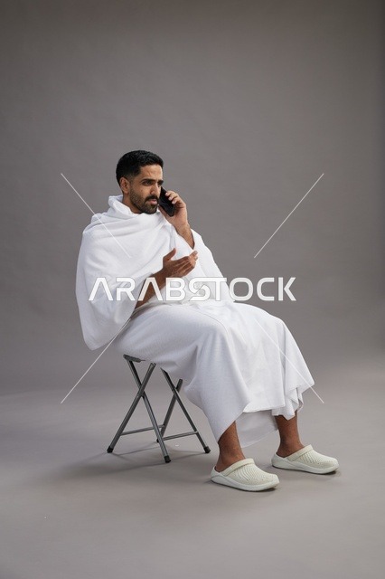 A portrait of a Saudi pilgrim in Ihram clothing using a mobile phone and sitting on a chair, preparing to perform the rituals of Hajj and Umrah, with a gray background.