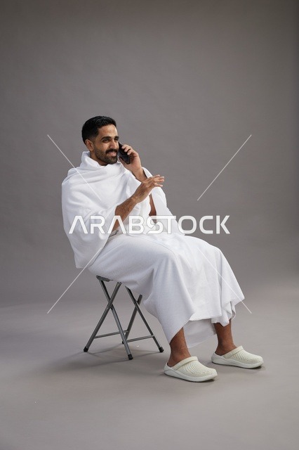 بورتريه لحاج سعودي بملابس الإحرام يستخدم الهاتف المحمول ويجلس على كرسي, الاستعداد لاداء مناسك الحج والعمره, خلفيه رماديه
