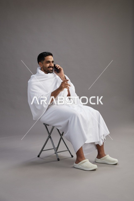 بورتريه لحاج سعودي بملابس الإحرام يستخدم الهاتف المحمول ويجلس على كرسي, الاستعداد لاداء مناسك الحج والعمره, خلفيه رماديه