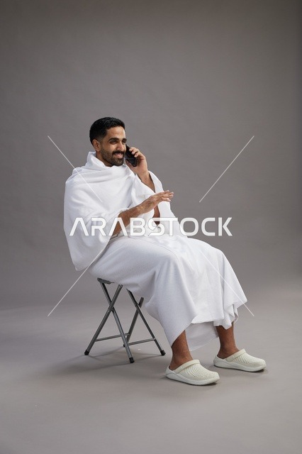 بورتريه لحاج سعودي بملابس الإحرام يستخدم الهاتف المحمول ويجلس على كرسي, الاستعداد لاداء مناسك الحج والعمره, خلفيه رماديه