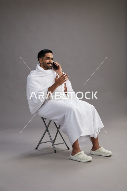 بورتريه لحاج سعودي بملابس الإحرام يستخدم الهاتف المحمول ويجلس على كرسي, الاستعداد لاداء مناسك الحج والعمره, خلفيه رماديه