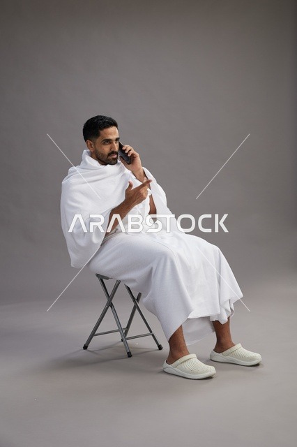 بورتريه لحاج سعودي بملابس الإحرام يستخدم الهاتف المحمول ويجلس على كرسي, الاستعداد لاداء مناسك الحج والعمره, خلفيه رماديه