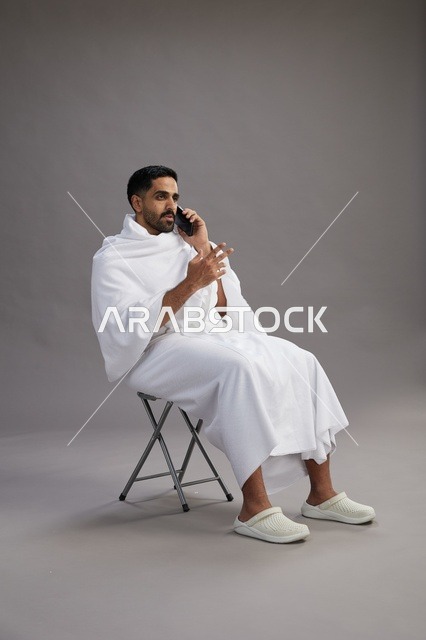 بورتريه لحاج سعودي بملابس الإحرام يستخدم الهاتف المحمول ويجلس على كرسي, الاستعداد لاداء مناسك الحج والعمره, خلفيه رماديه
