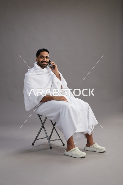 بورتريه لحاج سعودي بملابس الإحرام يستخدم الهاتف المحمول ويجلس على كرسي, الاستعداد لاداء مناسك الحج والعمره, خلفيه رماديه