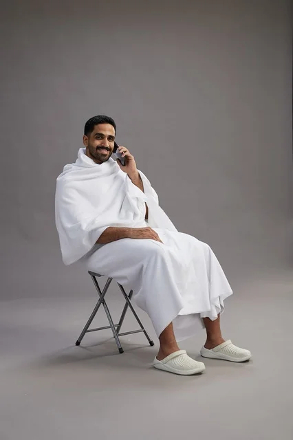 بورتريه لحاج سعودي بملابس الإحرام يستخدم الهاتف المحمول ويجلس على كرسي, الاستعداد لاداء مناسك الحج والعمره, خلفيه رماديه
