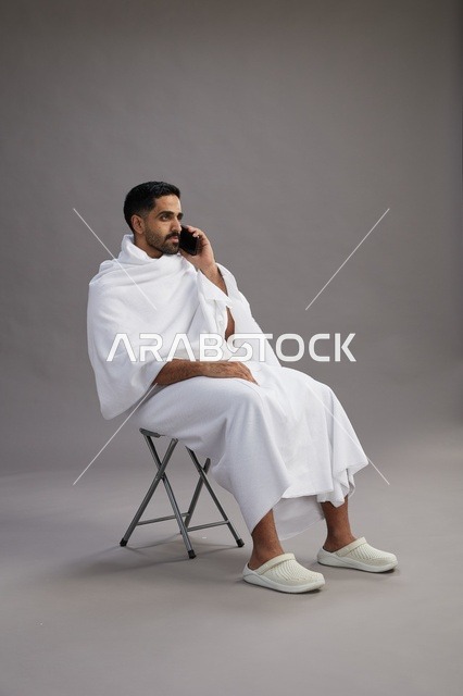 بورتريه لحاج سعودي بملابس الإحرام يستخدم الهاتف المحمول ويجلس على كرسي, الاستعداد لاداء مناسك الحج والعمره, خلفيه رماديه