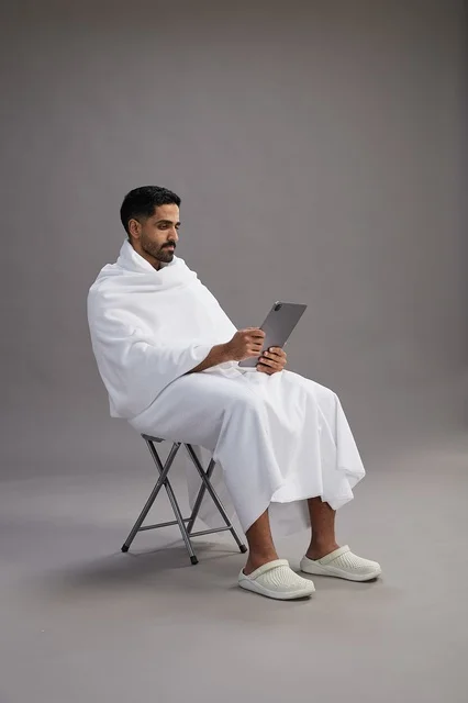 A portrait of a Saudi pilgrim in Ihram clothing holding a tablet and sitting on a chair, preparing to perform the rituals of Hajj and Umrah, with a gray background.