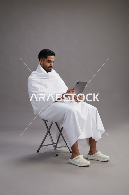 بورتريه لحاج سعودي بملابس الإحرام يحمل تابلت ويجلس على كرسي, الاستعداد لاداء مناسك الحج والعمره, خلفيه رماديه