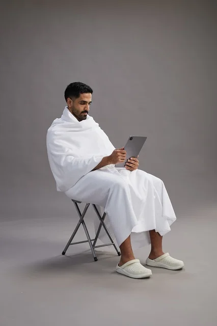 بورتريه لحاج سعودي بملابس الإحرام يحمل تابلت ويجلس على كرسي, الاستعداد لاداء مناسك الحج والعمره, خلفيه رماديه