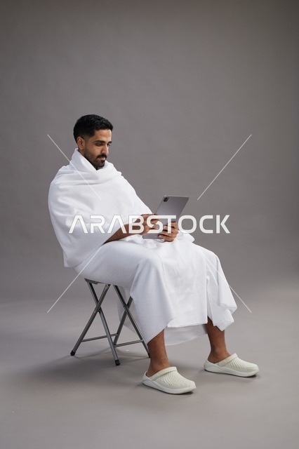 A portrait of a Saudi pilgrim in Ihram clothing holding a tablet and sitting on a chair, preparing to perform the rituals of Hajj and Umrah, with a gray background.