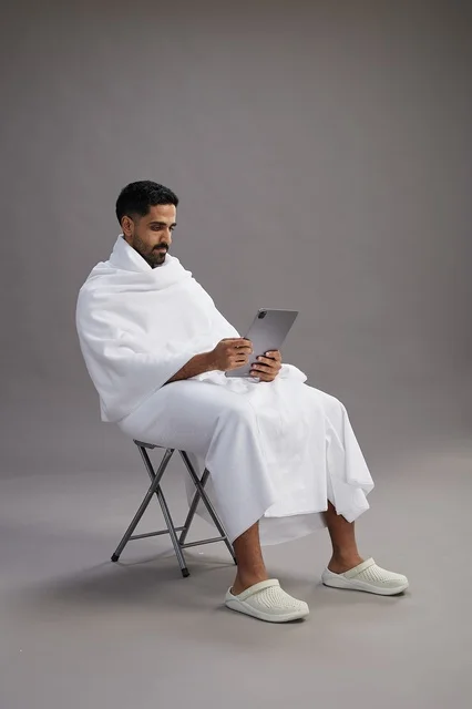 A portrait of a Saudi pilgrim in Ihram clothing holding a tablet and sitting on a chair, preparing to perform the rituals of Hajj and Umrah, with a gray background.