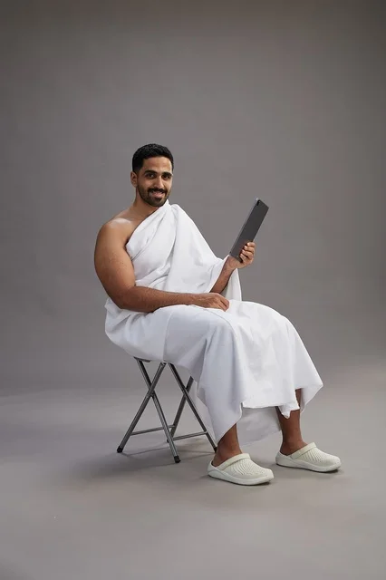 بورتريه لحاج سعودي بملابس الإحرام يحمل تابلت ويجلس على كرسي, الاستعداد لاداء مناسك الحج والعمره, خلفيه رماديه