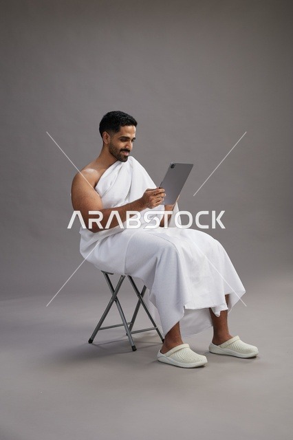 A portrait of a Saudi pilgrim in Ihram clothing holding a tablet and sitting on a chair, preparing to perform the rituals of Hajj and Umrah, with a gray background.