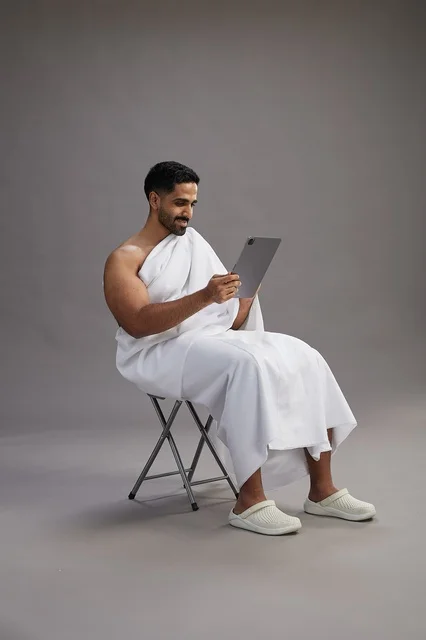 A portrait of a Saudi pilgrim in Ihram clothing holding a tablet and sitting on a chair, preparing to perform the rituals of Hajj and Umrah, with a gray background.