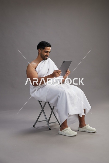 A portrait of a Saudi pilgrim in Ihram clothing holding a tablet and sitting on a chair, preparing to perform the rituals of Hajj and Umrah, with a gray background.