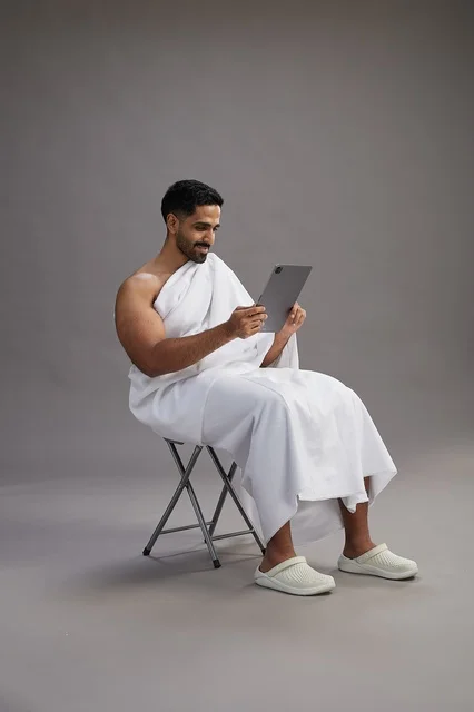 بورتريه لحاج سعودي بملابس الإحرام يحمل تابلت ويجلس على كرسي, الاستعداد لاداء مناسك الحج والعمره, خلفيه رماديه