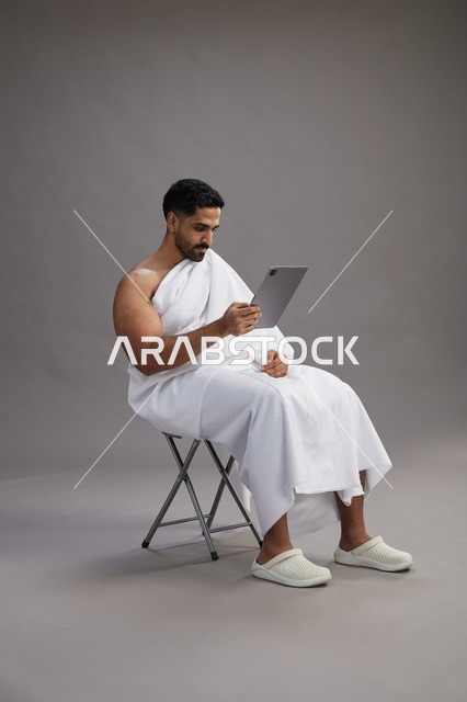 بورتريه لحاج سعودي بملابس الإحرام يحمل تابلت ويجلس على كرسي, الاستعداد لاداء مناسك الحج والعمره, خلفيه رماديه