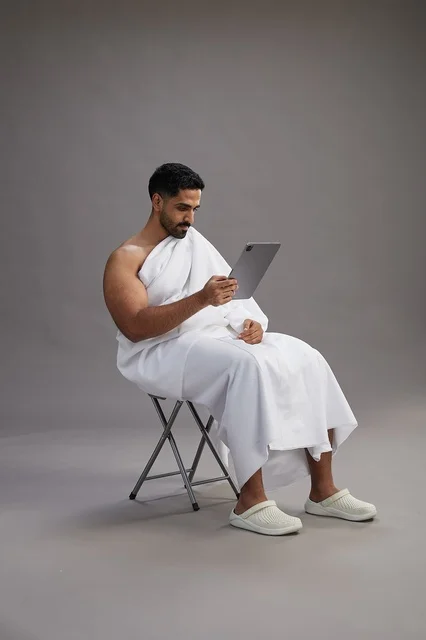 بورتريه لحاج سعودي بملابس الإحرام يحمل تابلت ويجلس على كرسي, الاستعداد لاداء مناسك الحج والعمره, خلفيه رماديه