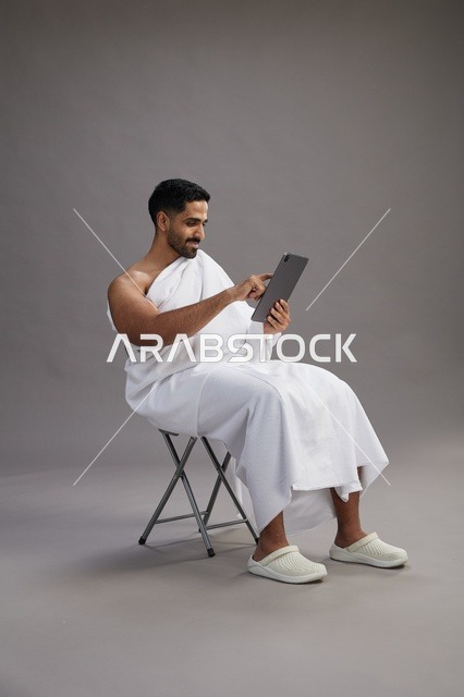 A portrait of a Saudi pilgrim in Ihram clothing holding a tablet and sitting on a chair, preparing to perform the rituals of Hajj and Umrah, with a gray background.